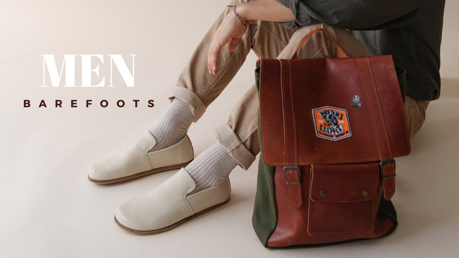 Man seated in Barefoots light slip-ons, beige cuffed pants, and brown leather bag with patches. Text 'Men Barefoots' in bold white font.