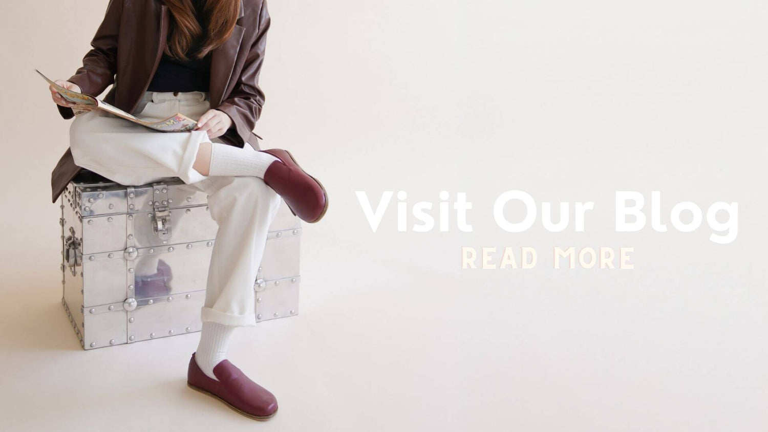 A young woman reading a magazine. she is dressed in a brown leather jacket, white pants, and burgundy barefoot loafers, against a minimalist background.