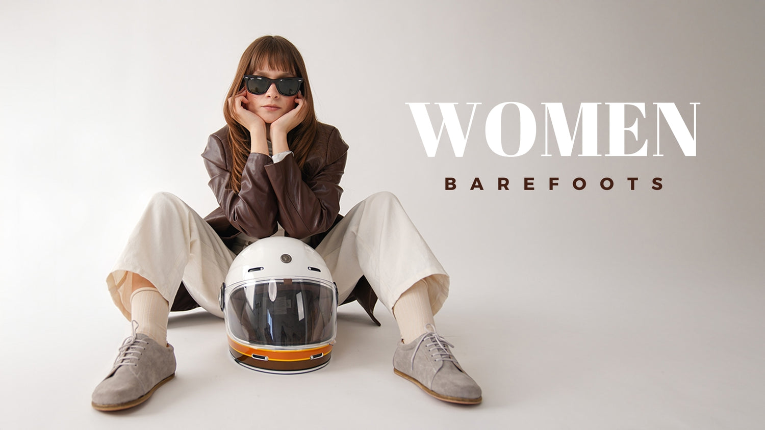Woman in casual outfit wearing gray barefoot shoes, sitting with a motorcycle helmet, representing women's barefoot footwear collection.