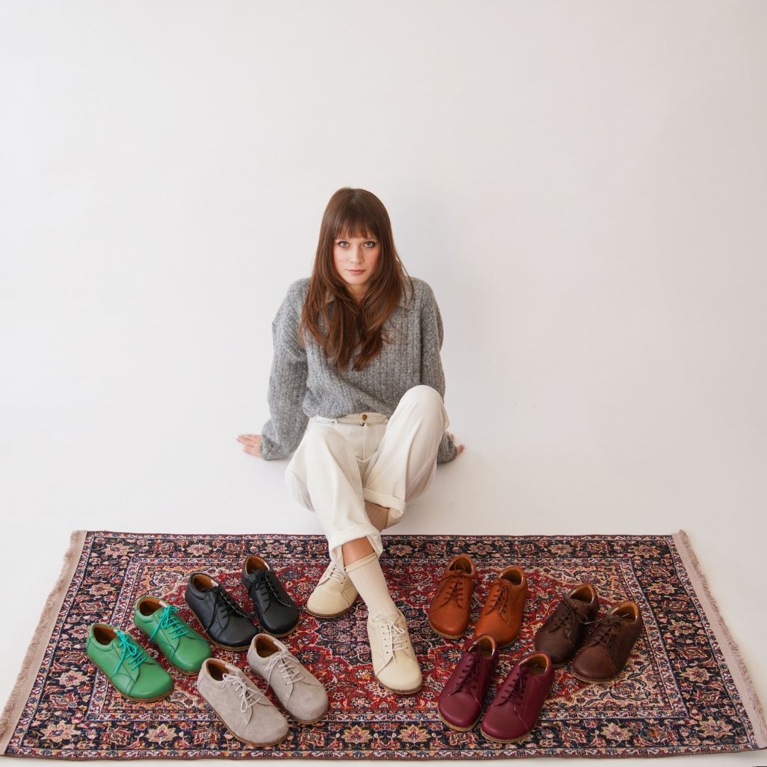Woman in grey sweater and white pants sitting on a rug with various colorful shoes.