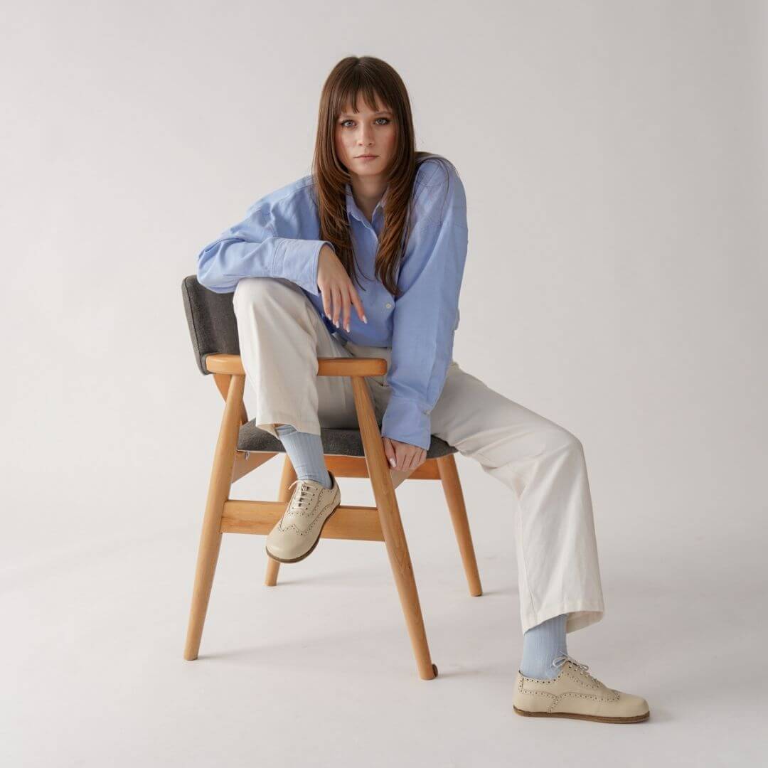 Woman in a blue shirt and cream pants, sitting on a wooden chair in beige shoes.