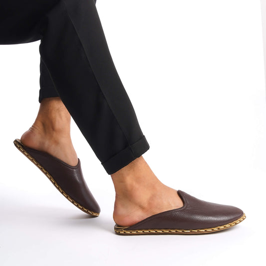 Person lifting foot while wearing dark brown leather slippers with natural leather soles, zero drop design, and edge stitching.