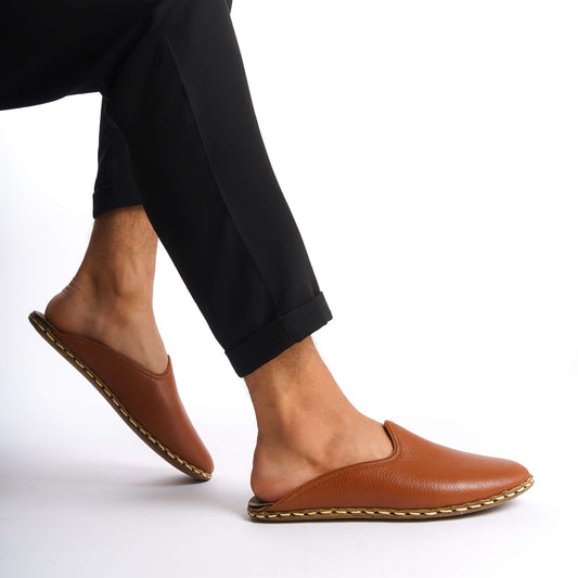 Person lifting foot while wearing brown leather slippers with natural leather soles, zero drop design, and edge stitching.