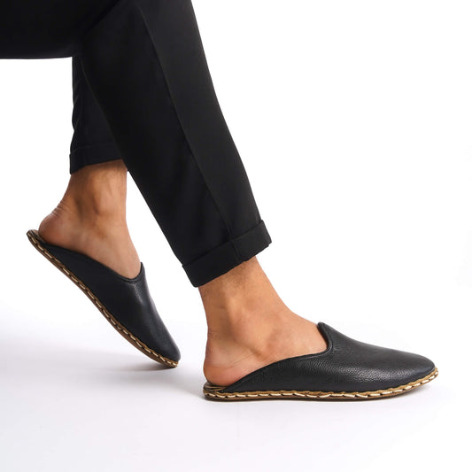 Person lifting foot while wearing black leather slippers with natural leather soles, zero drop design, and edge stitching.