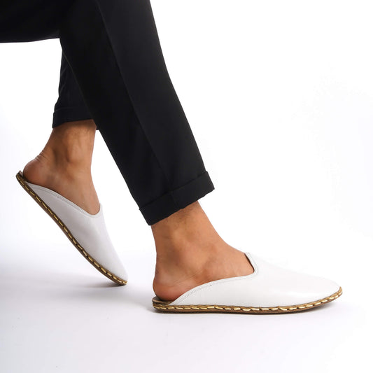 Person lifting foot while wearing white leather slippers with natural leather soles, zero drop design, and edge stitching.