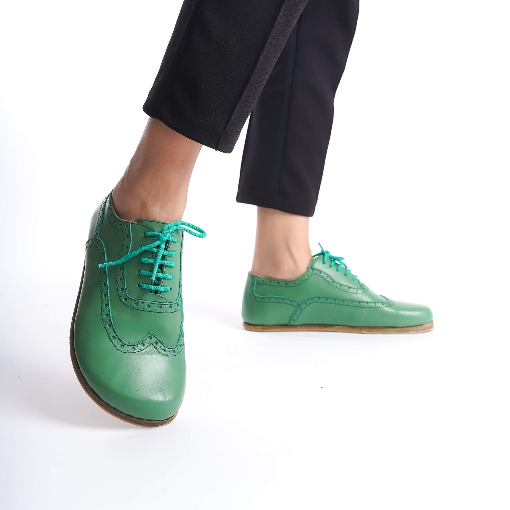 Model walking in green Doris Leather Barefoot Women Oxfords with teal laces and black pants.