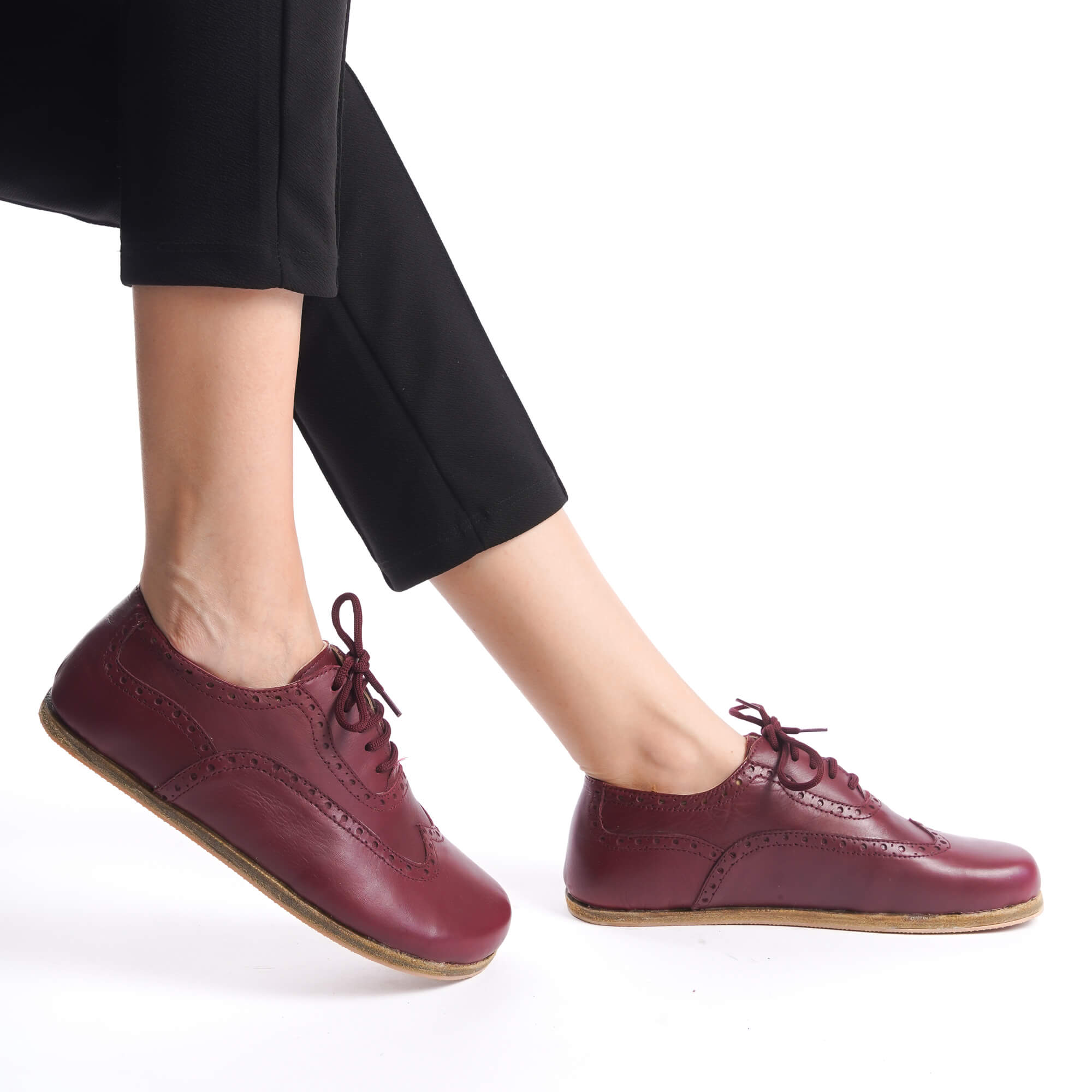A person sitting with legs crossed, showcasing Doris leather barefoot women Oxfords in Burgundy against a white background.