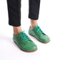Green Lydia leather barefoot women's sneakers with turquoise laces, worn by a model in black pants, showcasing the shoes' comfortable and flexible design.