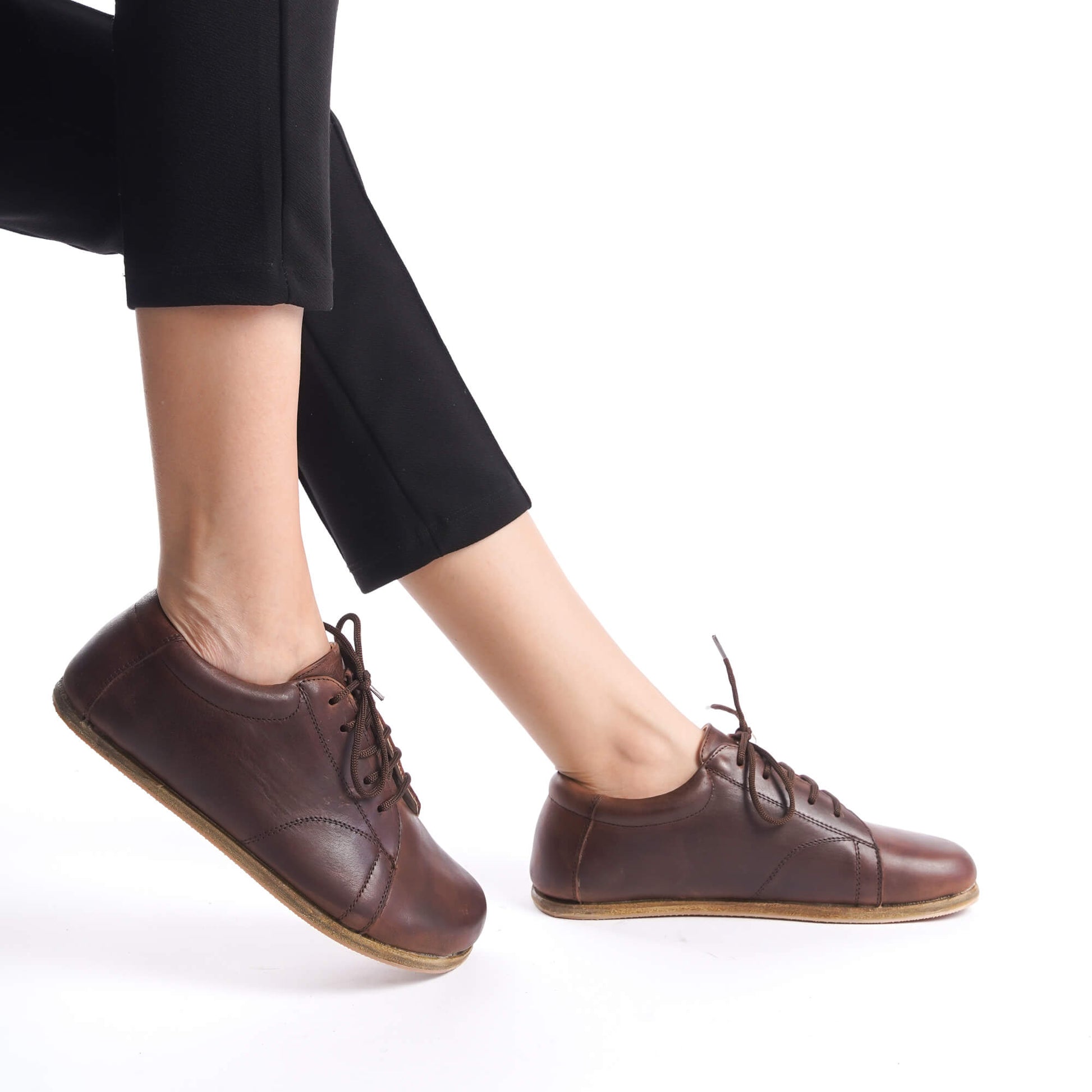 Side view of Lydia Leather Barefoot Women Sneakers in brown, worn by a model with black pants, emphasizing comfort and style.