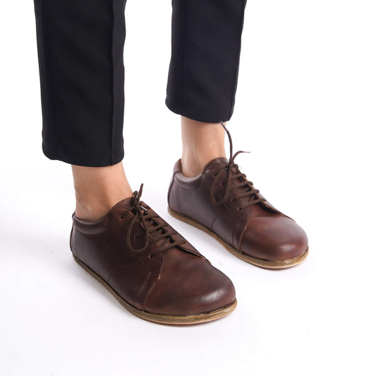 Close-up view of Lydia Leather Barefoot Women Sneakers in brown, worn by a model with black pants, showcasing the natural fit and leather material.