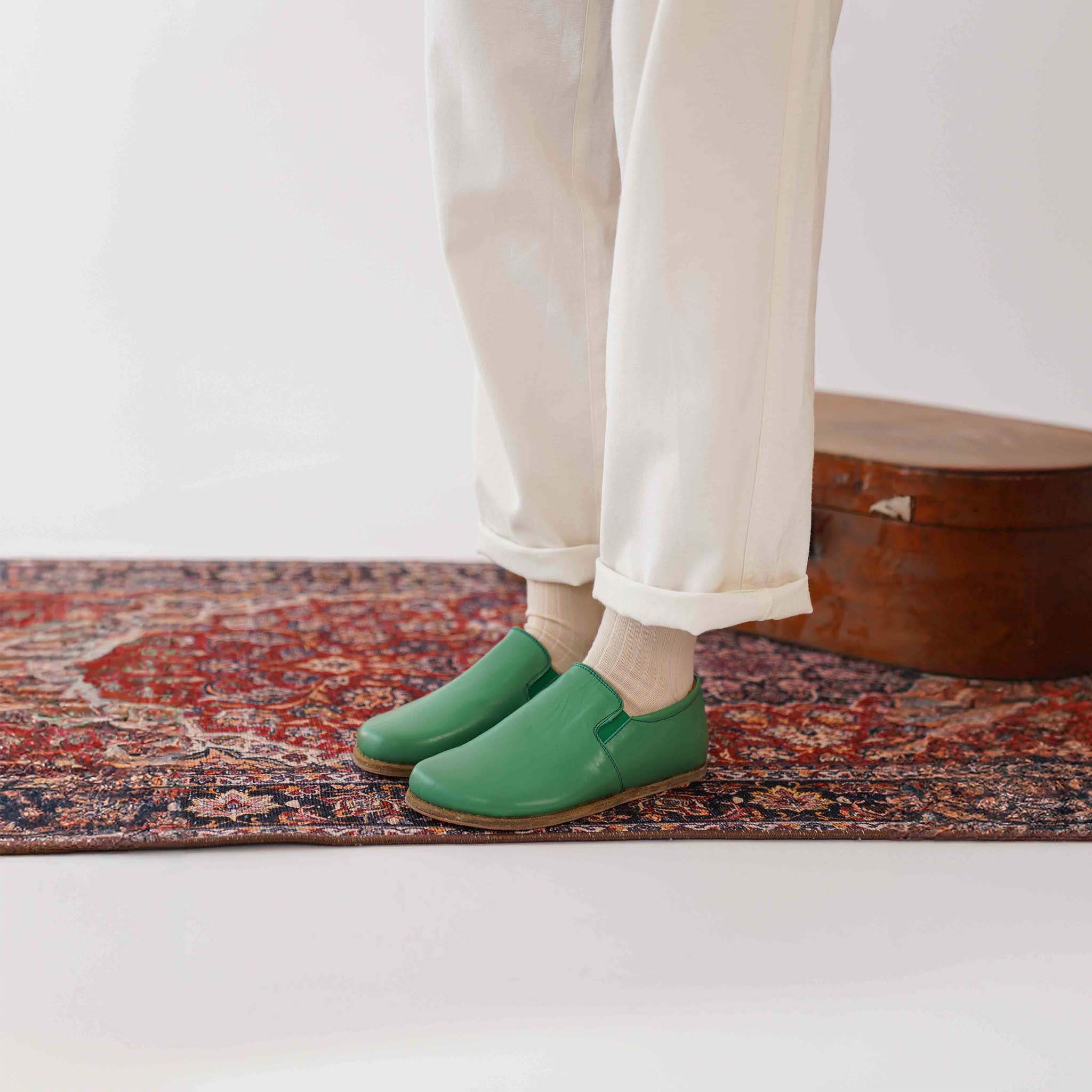 Model standing on a vintage suitcase in green leather loafers. Experience minimalist footwear at Pelanir.