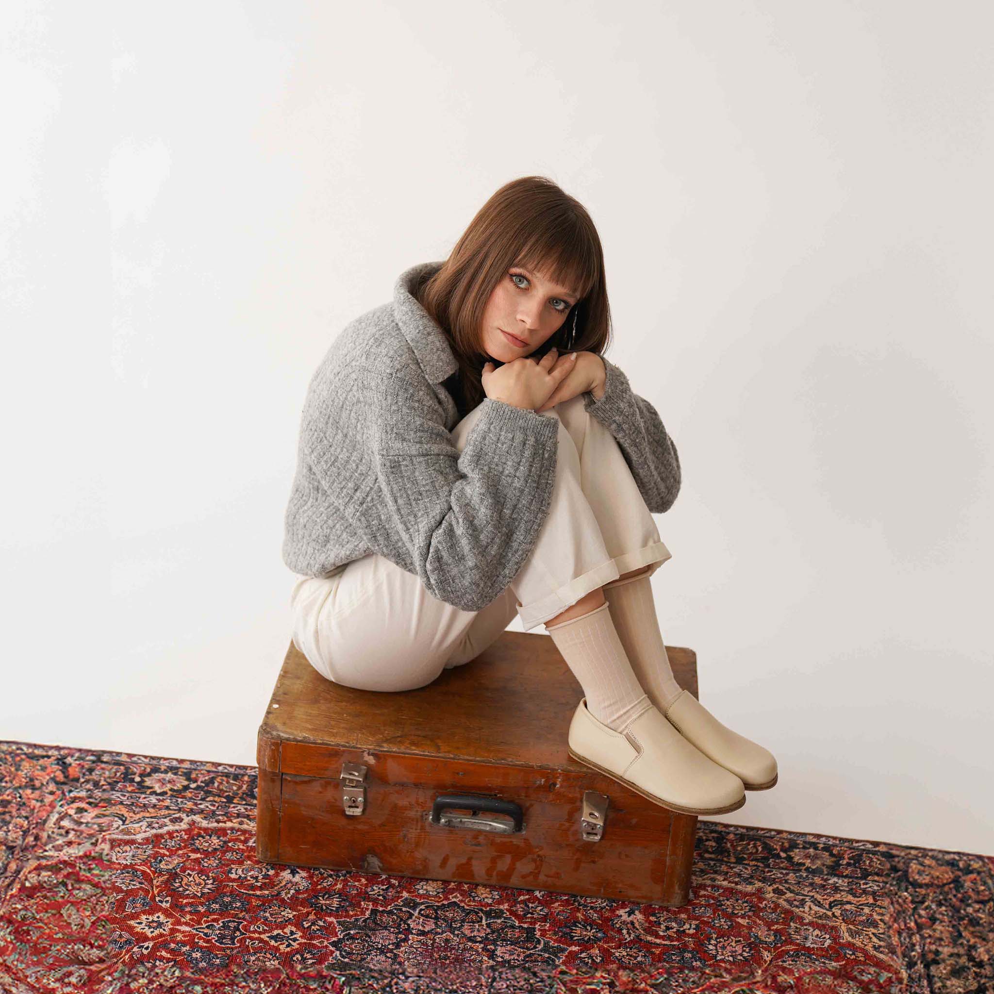 Model posing with Ionia beige loafers on a vintage suitcase - Step into comfort with Ionia Leather Barefoot Women Loafers in beige.
