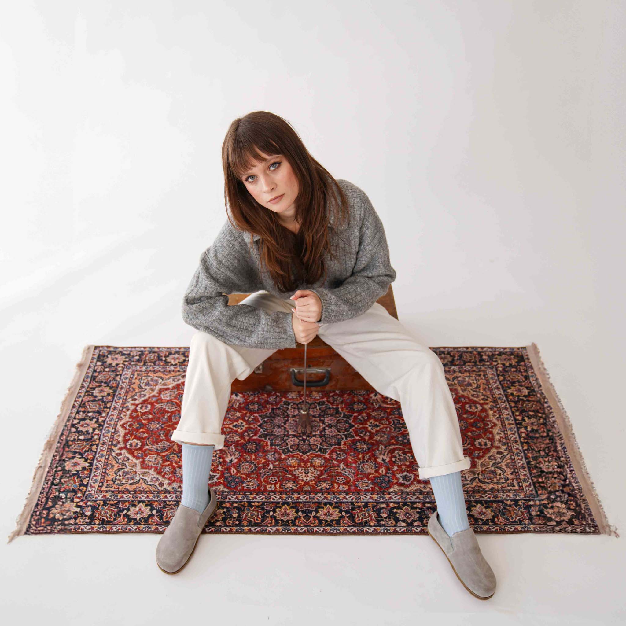 Model sitting on Turkish rug wearing gray suede Ionia leather loafers and white pants for a chic look.