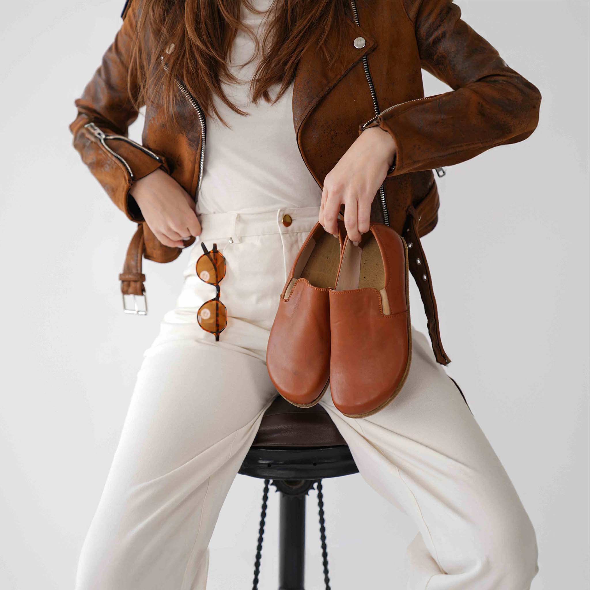 Model holding a pair of tan brown Ionia leather barefoot loafers, paired with a brown jacket and white pants for a chic look.