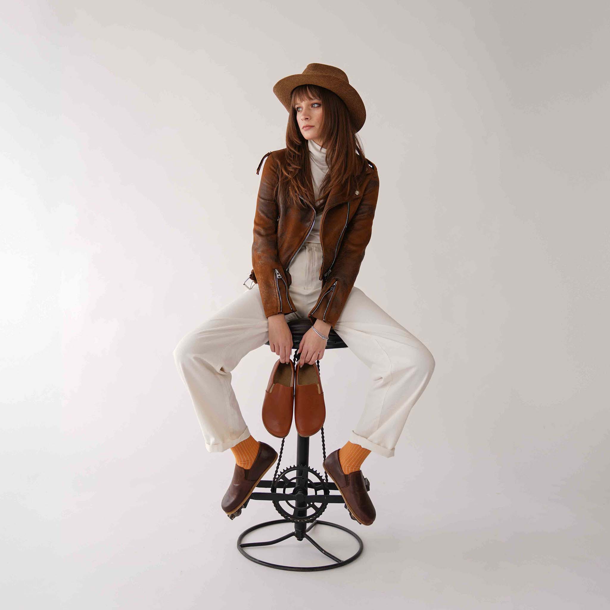 Model sitting on a stool, holding a pair of Ionia Leather Barefoot Women Loafers in brown, with matching brown jacket.