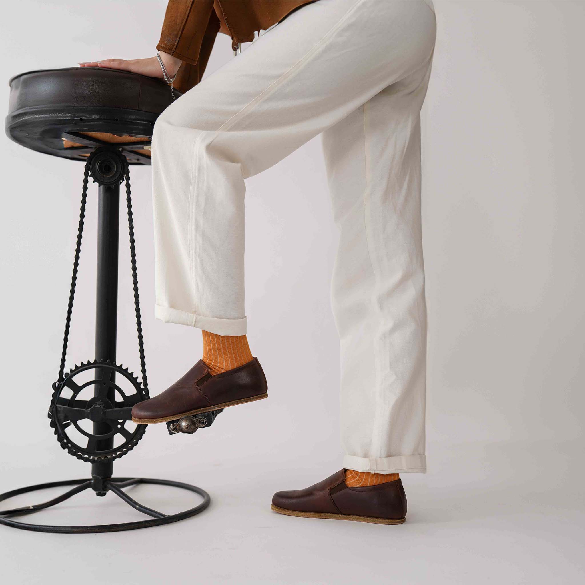 Model standing beside a unique stool, wearing Ionia Leather Barefoot Women Loafers in brown with orange socks.