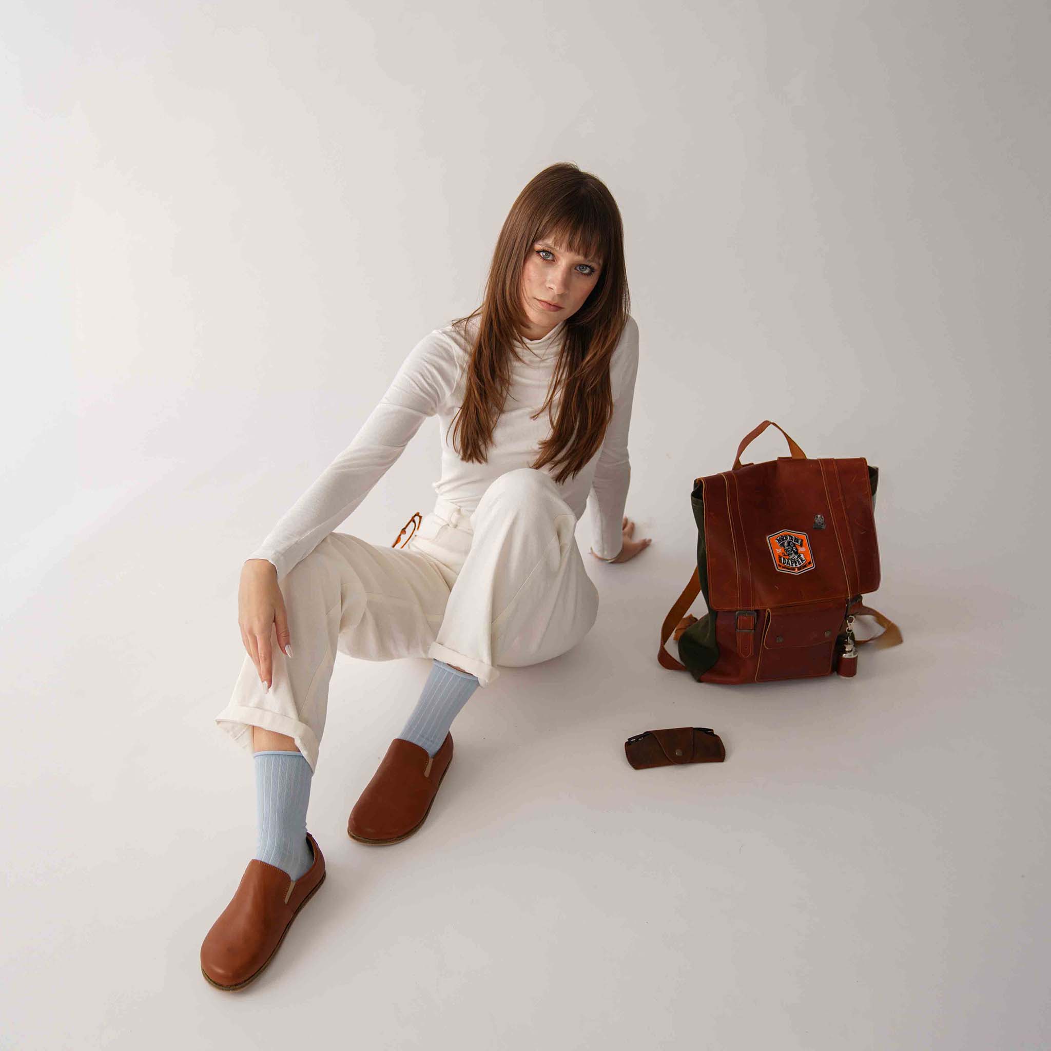 Model wearing tan brown Ionia leather barefoot loafers, paired with a white outfit and sitting beside a leather backpack.