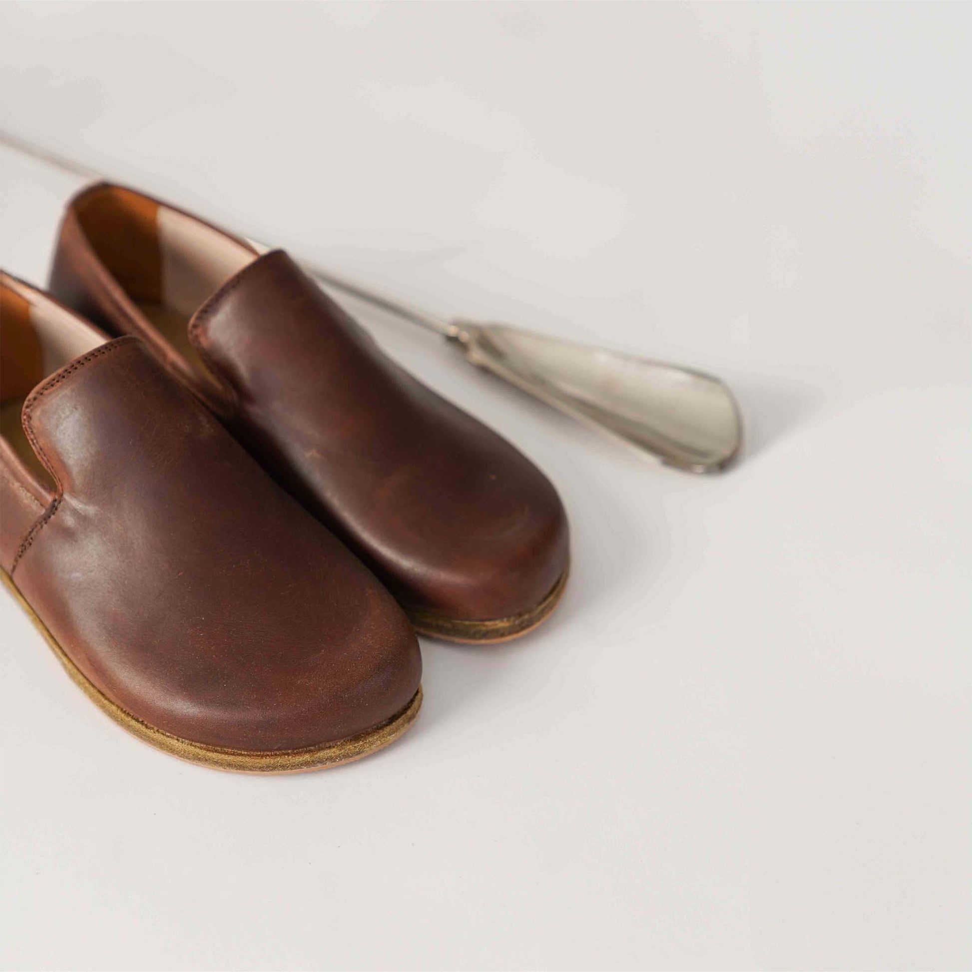 Pair of Aeolia Brown Women Loafers next to a metal shoehorn, showcasing the shoe's high-quality leather and minimalist design.