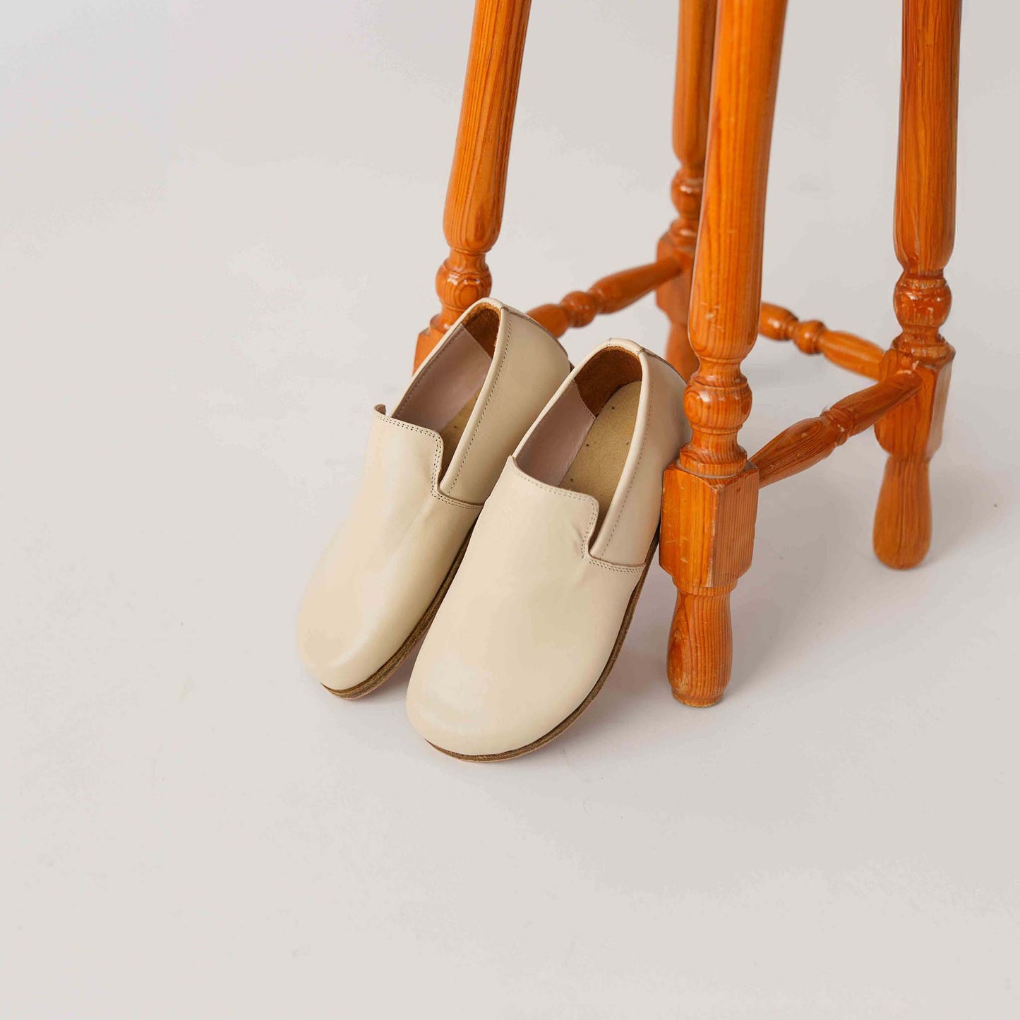 Pair of Aeolia Beige Women Loafers leaning against a wooden stool, highlighting the minimalist design and smooth leather finish.