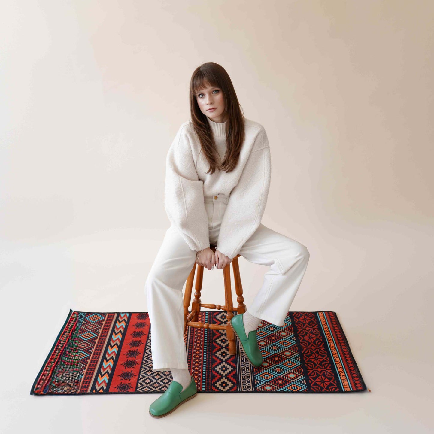 Model wearing Aeolia green leather barefoot women loafers, paired with white pants, seated on a wooden stool on a colorful patterned rug.