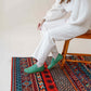 Model wearing Aeolia green leather barefoot women loafers, paired with white pants, seated on a wooden chair on a colorful rug.