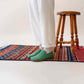 Aeolia green leather barefoot women loafers, paired with white pants and socks, standing on a colorful rug near a wooden stool.