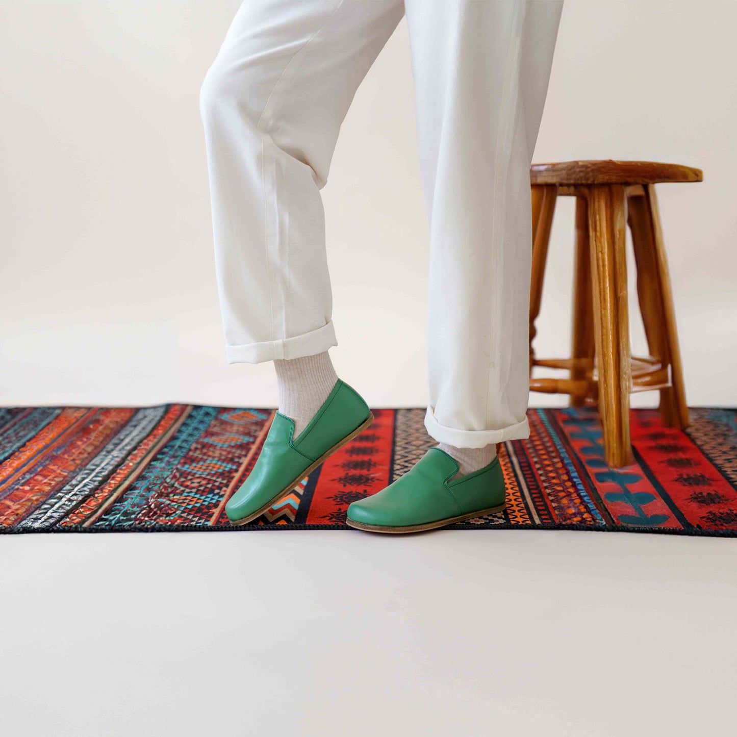 Aeolia green leather barefoot women loafers, paired with white pants and socks, standing on a colorful rug near a wooden stool.