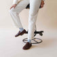 Model sitting on a modern stool, wearing white pants, light blue socks, and Aeolia Brown Women Loafers, showcasing the shoe's stylish and comfortable fit.