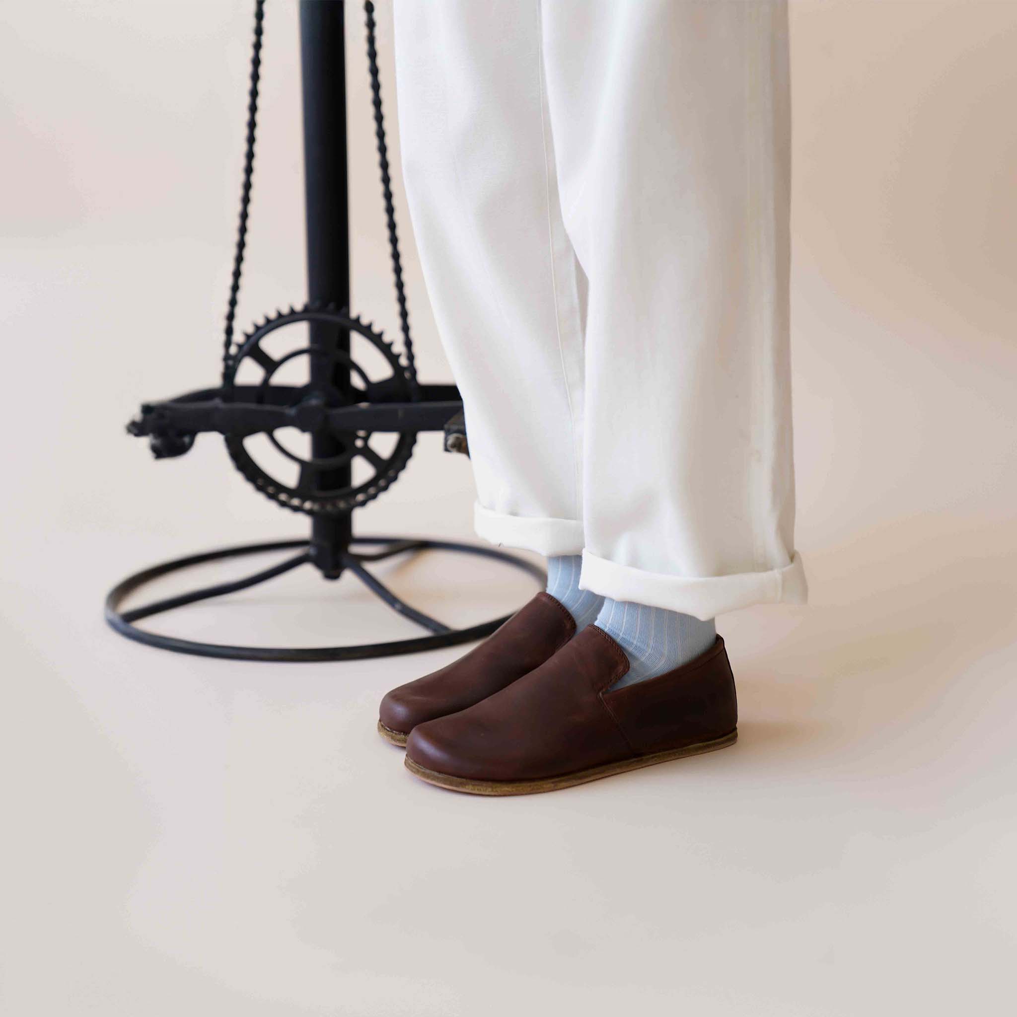 Model wearing white pants and Aeolia Brown Women Loafers with light blue socks, standing by a modern stool, showcasing the loafer's stylish fit.