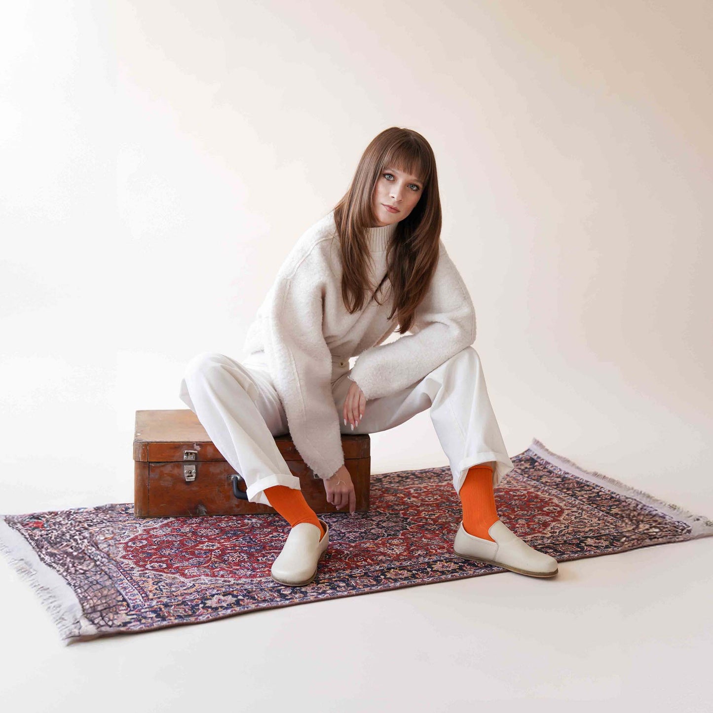 Model sitting on a patterned rug, wearing Aeolia Leather Barefoot Women Loafers in Beige with orange socks and a white outfit, posing casually.