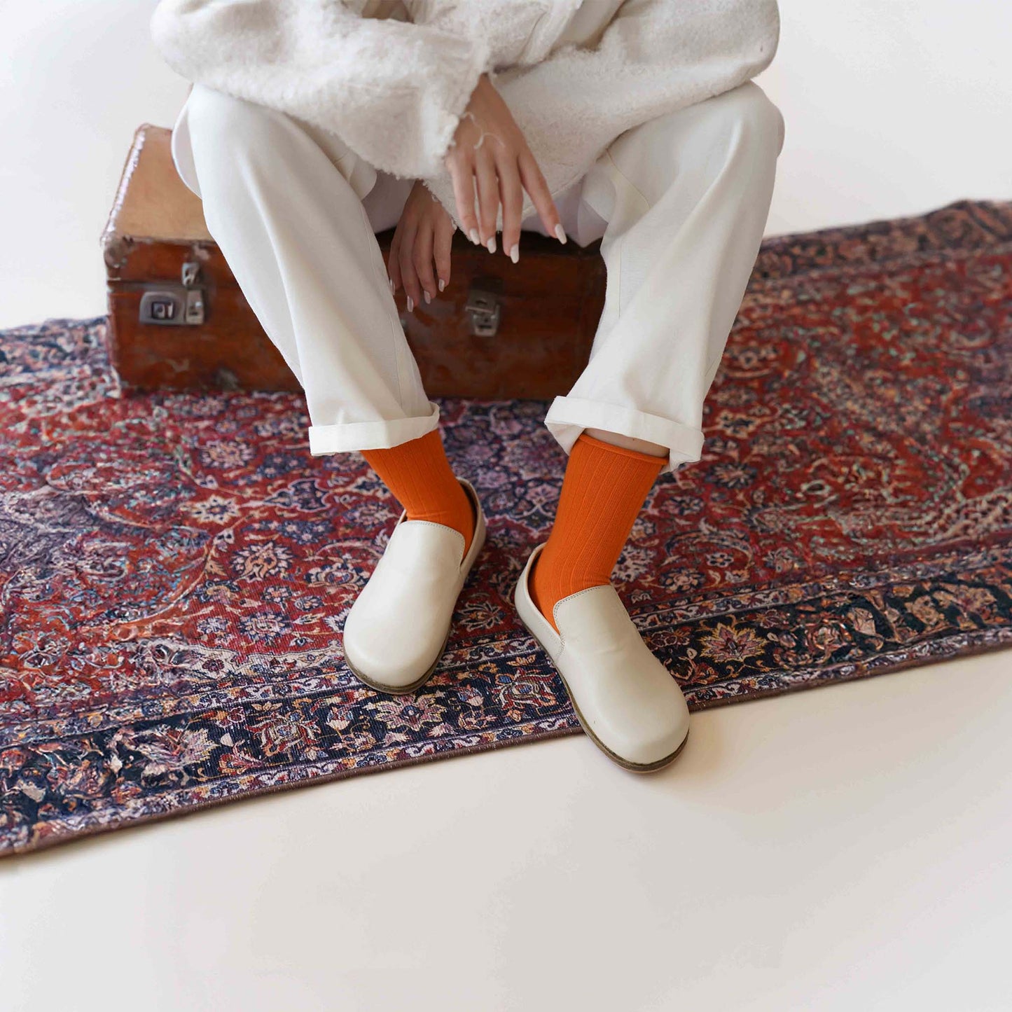 Model sitting on a suitcase with Aeolia Beige Loafers, orange socks, and white outfit on a patterned rug - stylish fit