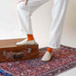 Model's foot on a suitcase, wearing orange socks and Aeolia Beige Loafers, standing on a patterned rug, showcasing the stylish fit.