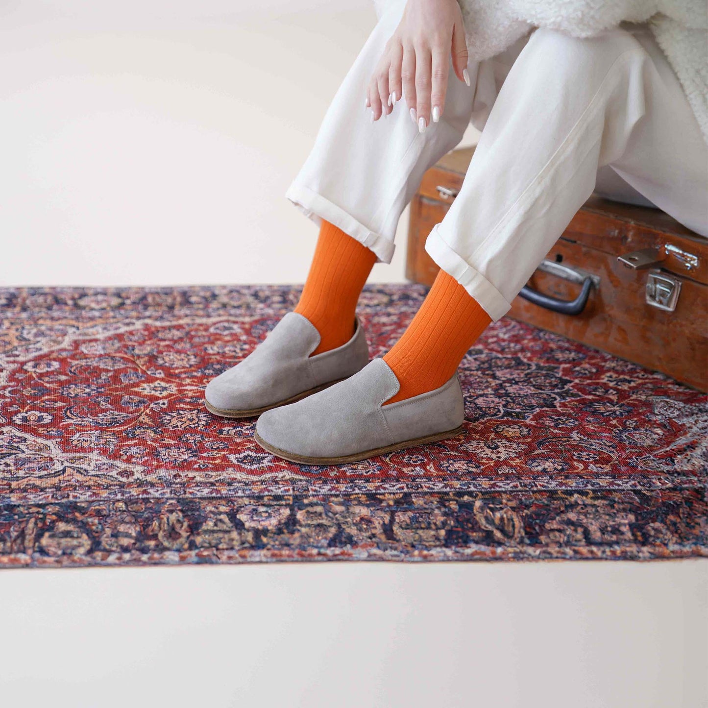 Aeolia gray suede barefoot women loafers, paired with white pants and orange socks, on a patterned rug by a vintage suitcase.