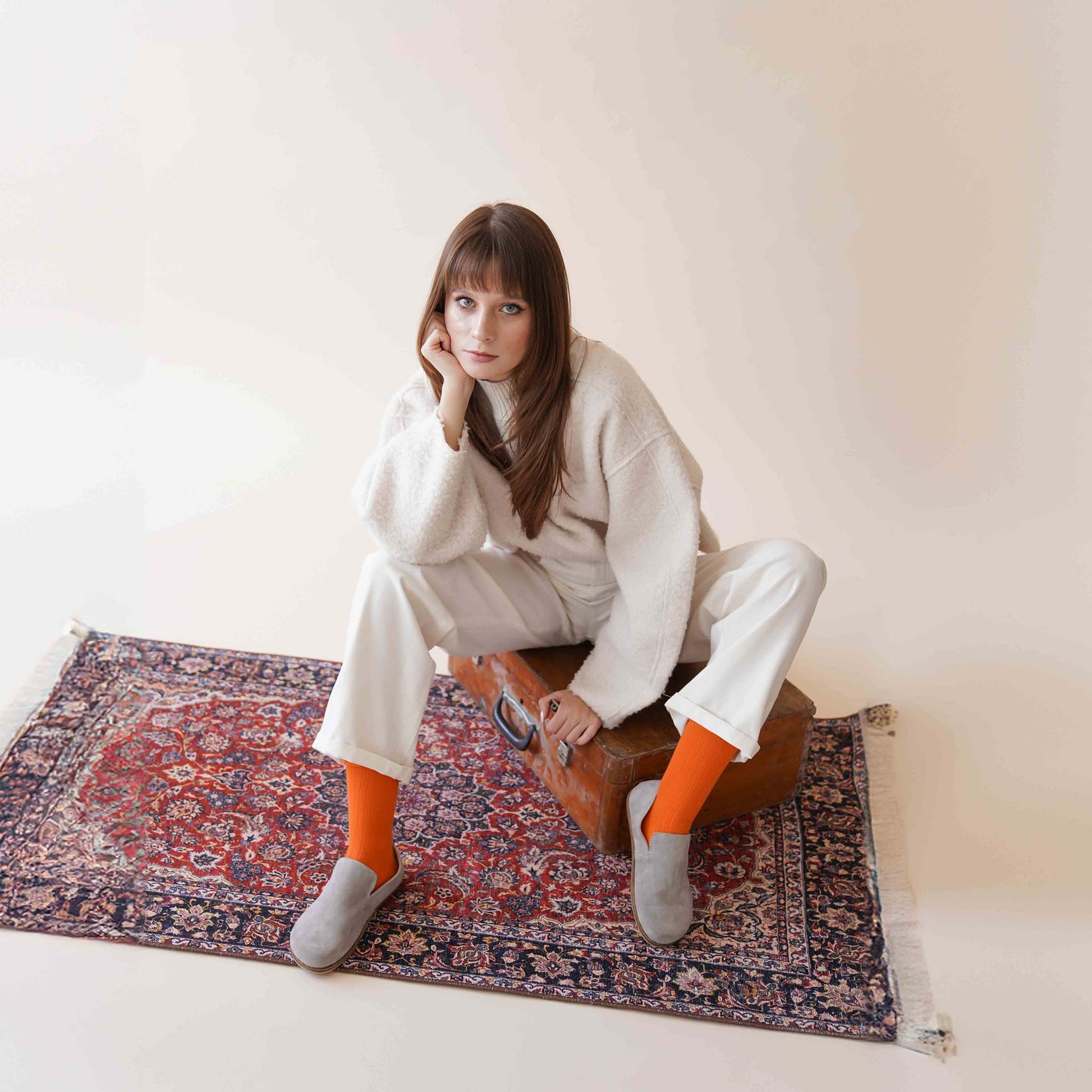 Model wearing Aeolia gray suede barefoot loafers, white pants, orange socks, seated on a vintage suitcase on a patterned rug.