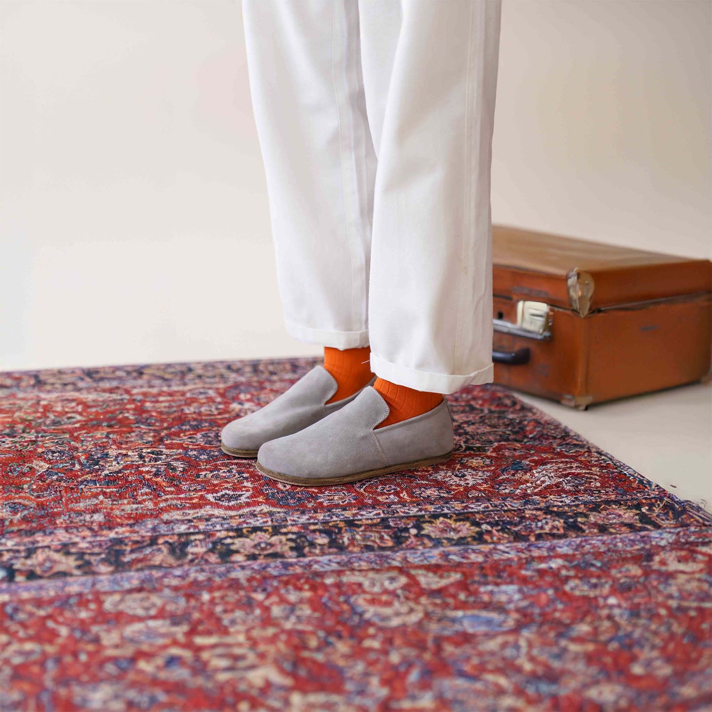 Aeolia gray suede barefoot women loafers, paired with white pants and orange socks, on a patterned rug beside a vintage suitcase.