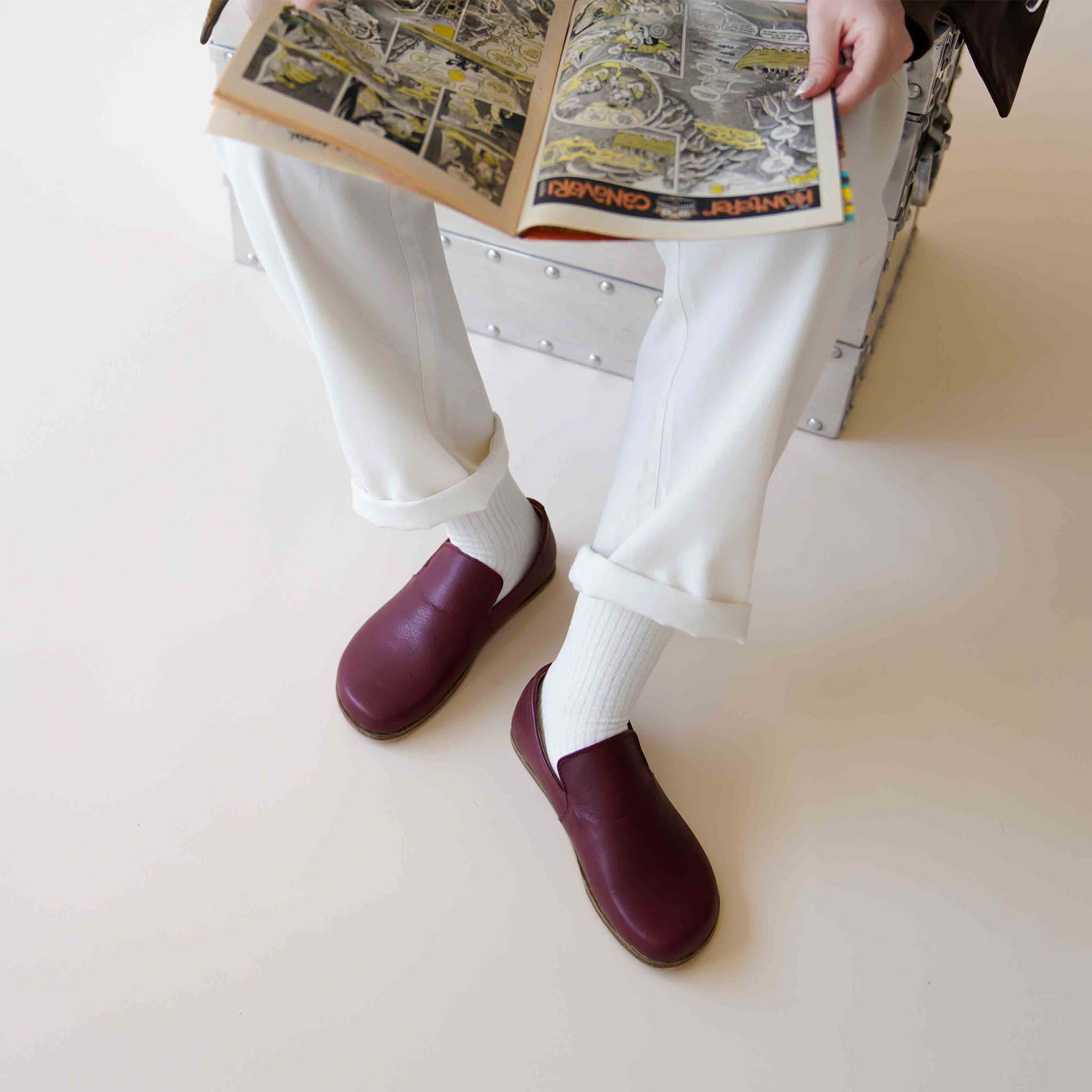 Aeolia Leather Barefoot Women Loafers in Burgundy, paired with white pants and socks, reading a comic book.
