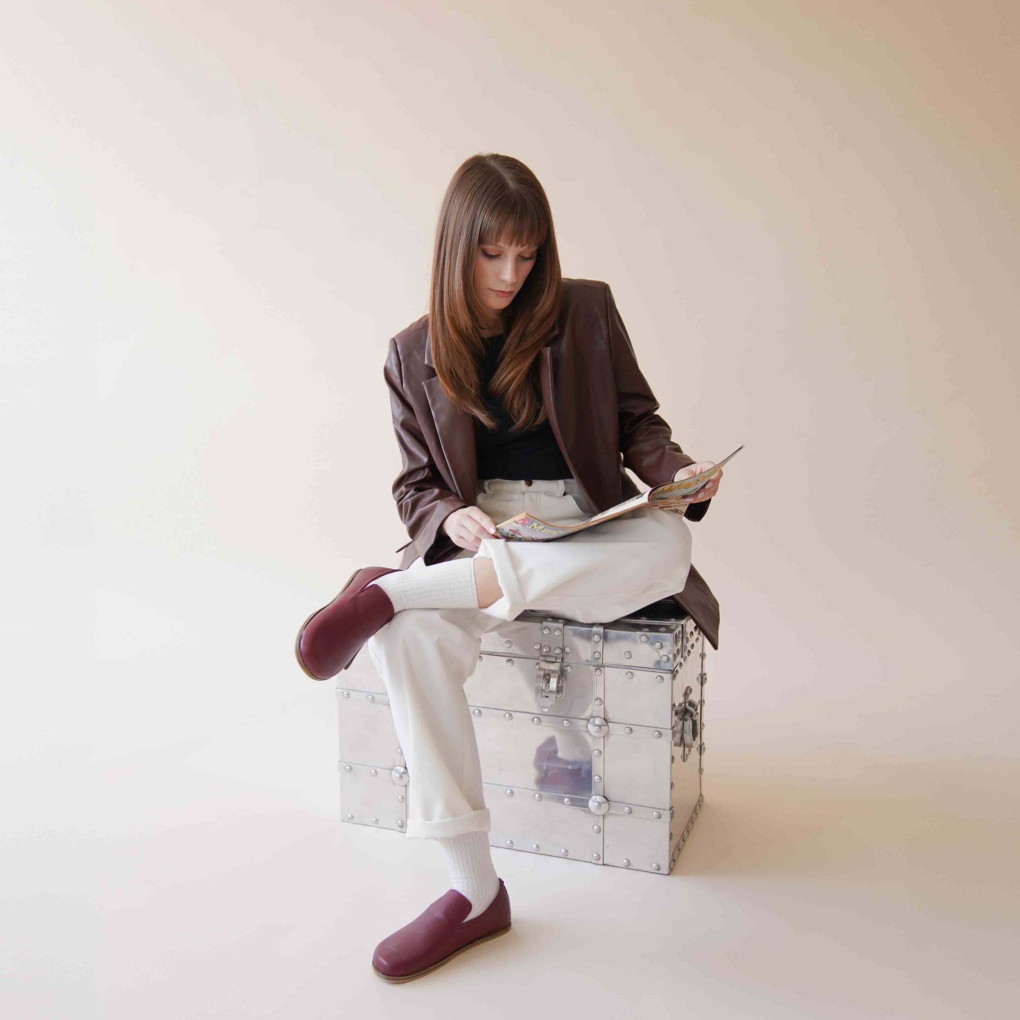 Model wearing Aeolia Leather Barefoot Women Loafers in Burgundy, seated on a metal chest, dressed in a brown jacket and white pants.