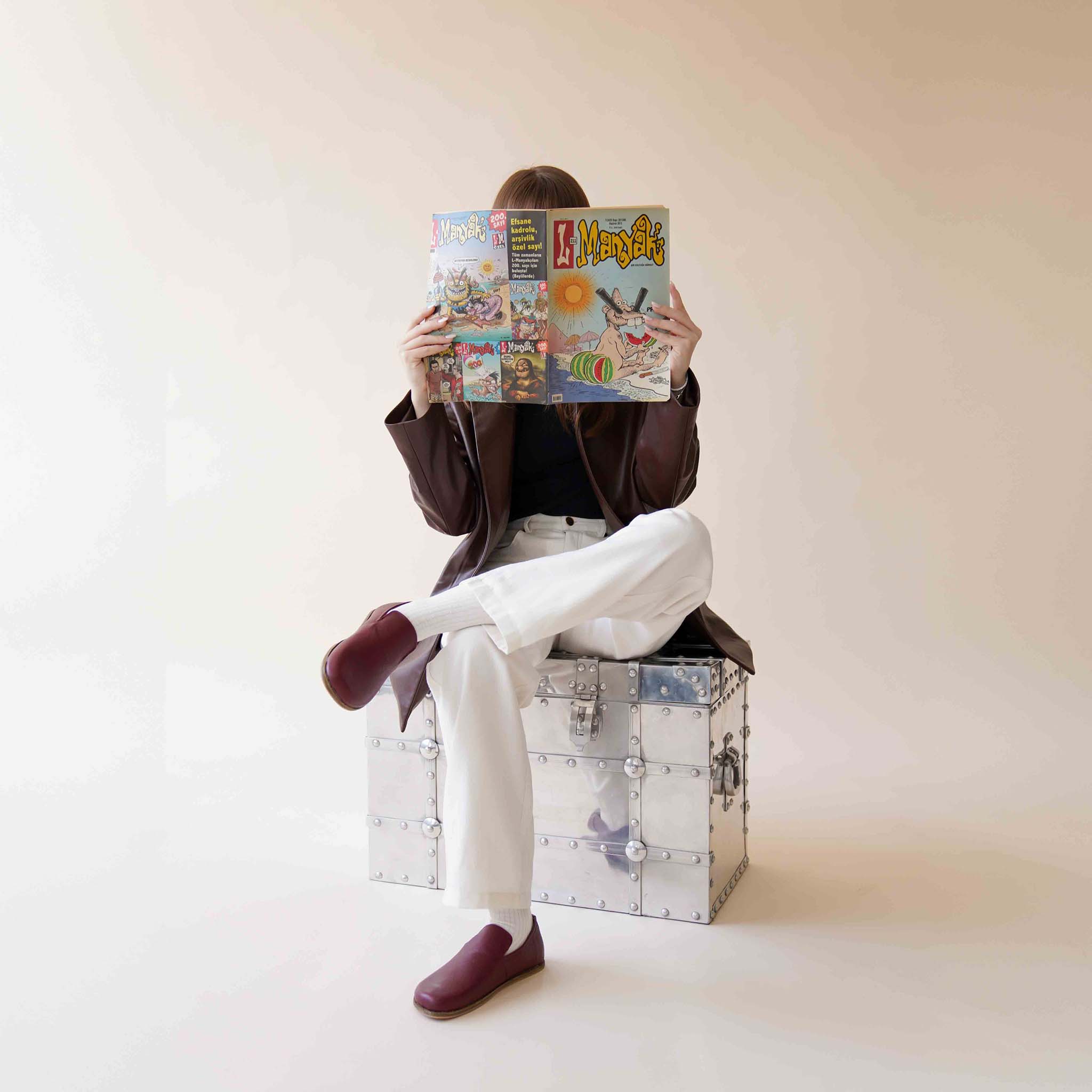 Model wearing Aeolia Leather Barefoot Women Loafers in Burgundy, seated on a metal chest, reading a comic book.