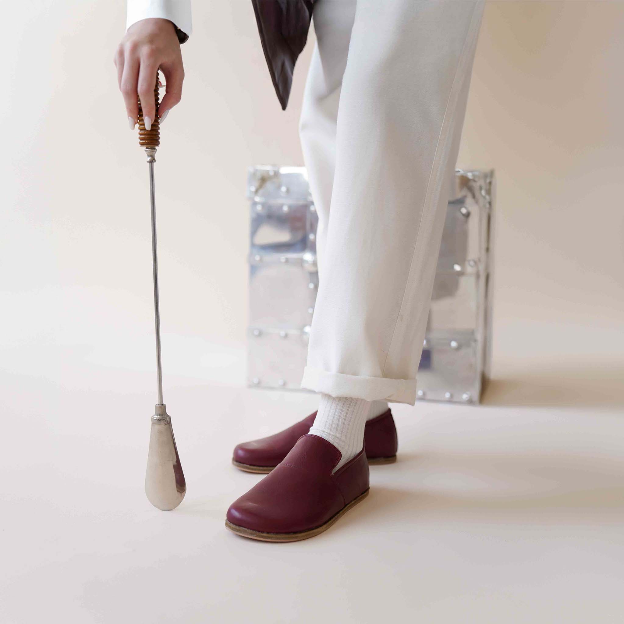 Wearing Aeolia Leather Barefoot Women Loafers in Burgundy, using a shoehorn, with white pants and socks.