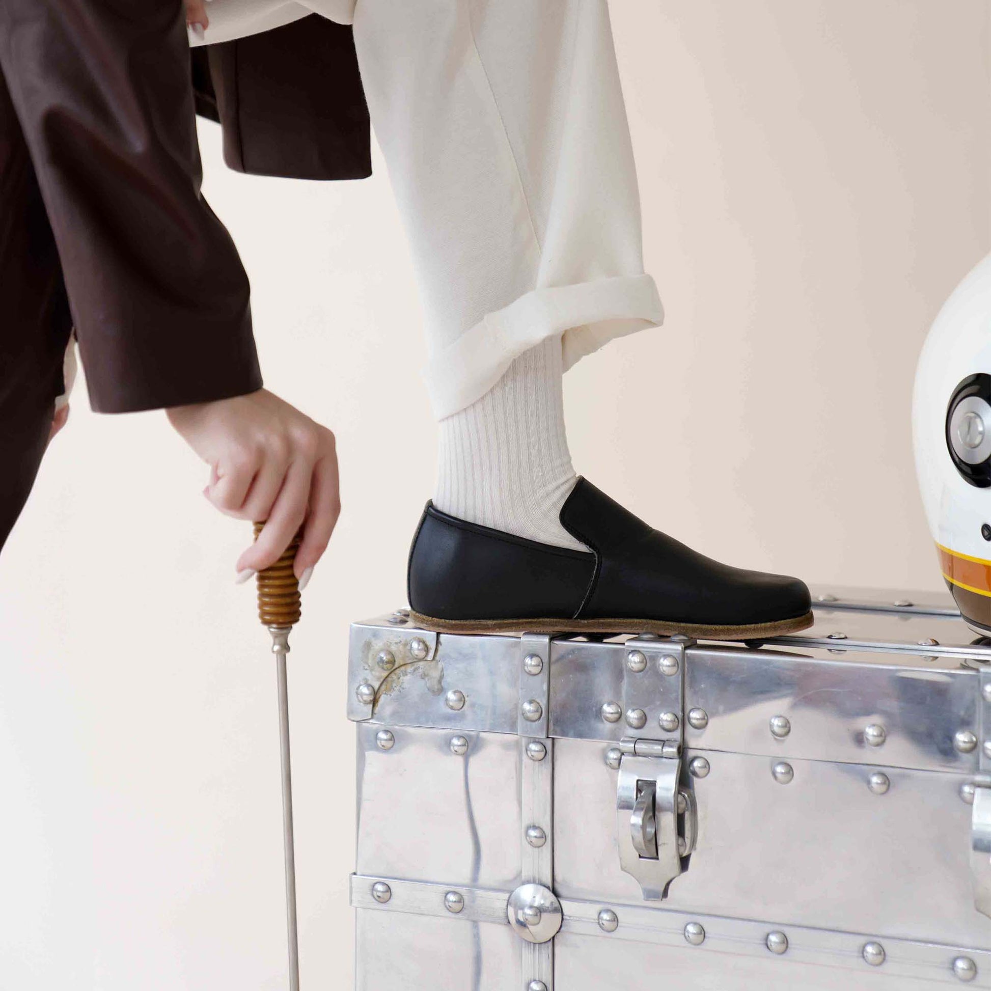 Close-up of Model's Foot Resting on a Metal Chest, Wearing Aeolia Leather Barefoot Women Loafers in Black - Showcasing the Shoe's Elegant and Minimalist Design.