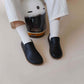 Model Sitting on the Floor Wearing Aeolia Leather Barefoot Women Loafers in Black and White Socks, with a White Helmet Beside Them - Showcasing the Loafer's Comfortable Fit and Minimalist Design.