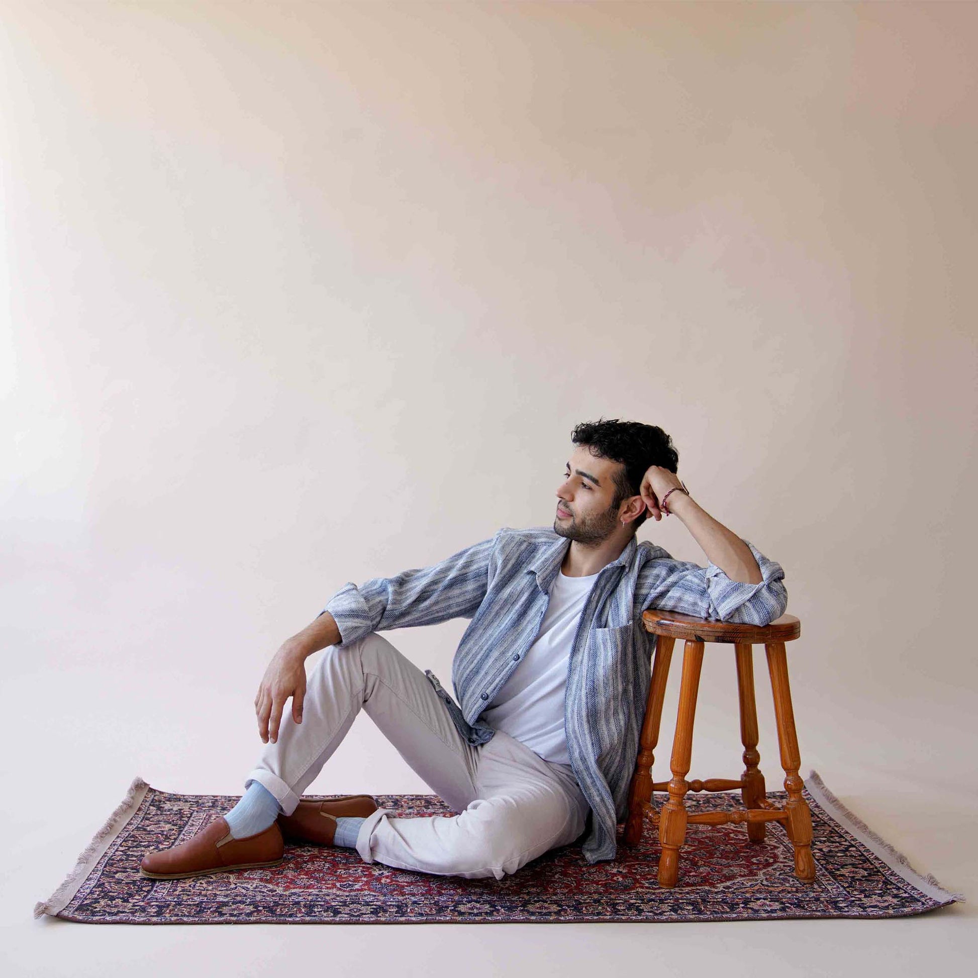 Model sitting on a rug, wearing tan brown Ionia leather barefoot loafers with white pants and a striped shirt, creating a relaxed and fashionable look. Discover more at pelanir.com!