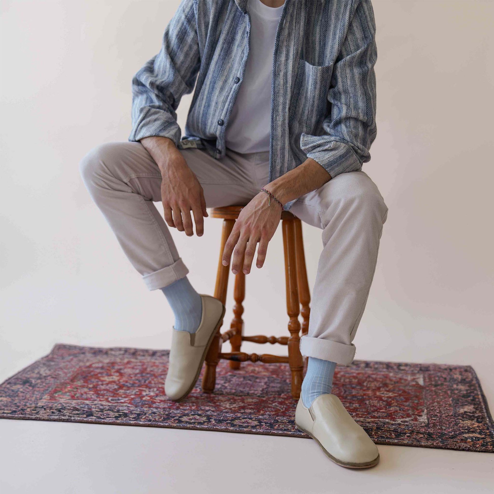 Model wearing Ionia Leather Barefoot Men Loafers in beige, paired with light-colored pants and a striped shirt, sitting on a stool.