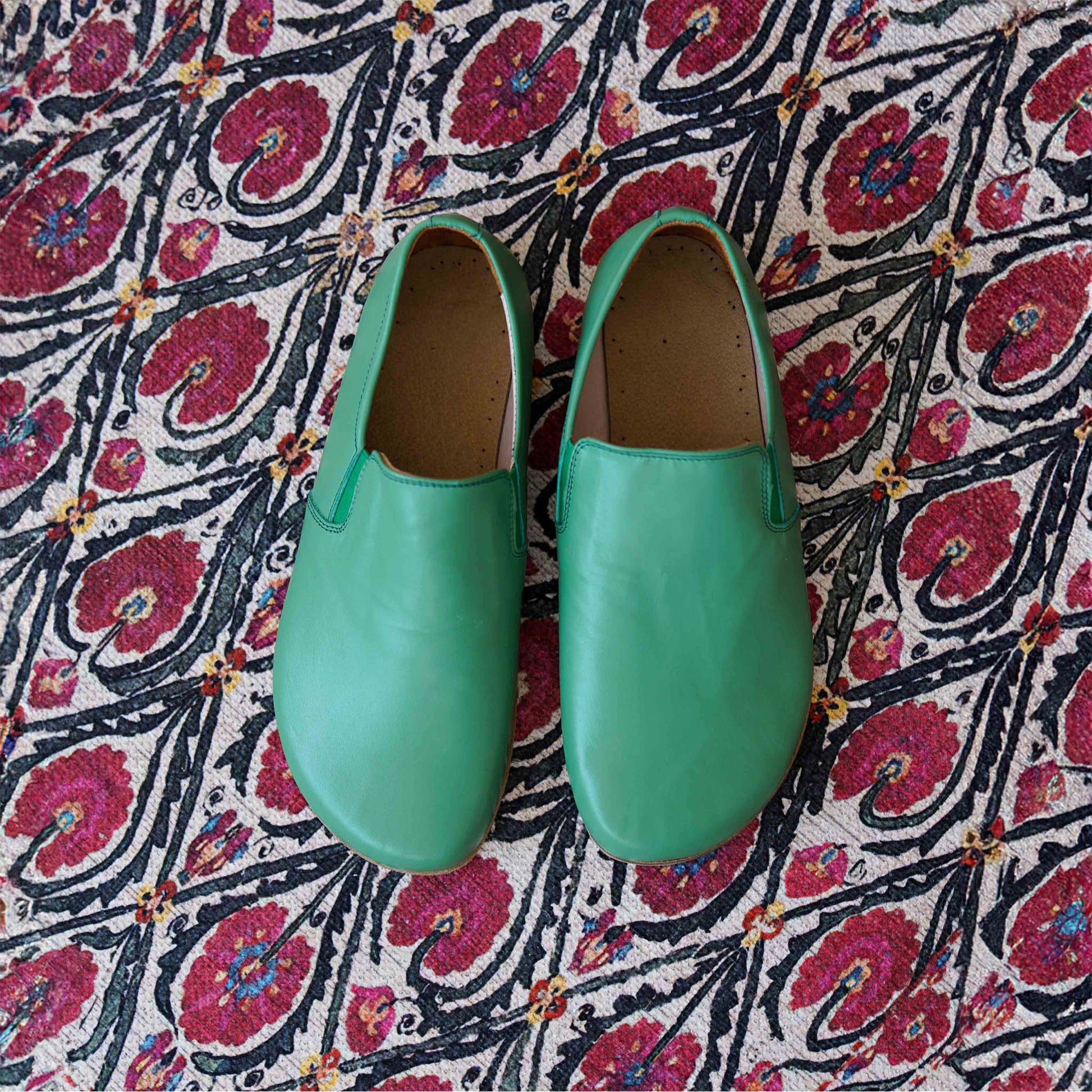 Green Ionia leather barefoot loafers on a floral patterned rug. Visit pelanir.com to buy now!