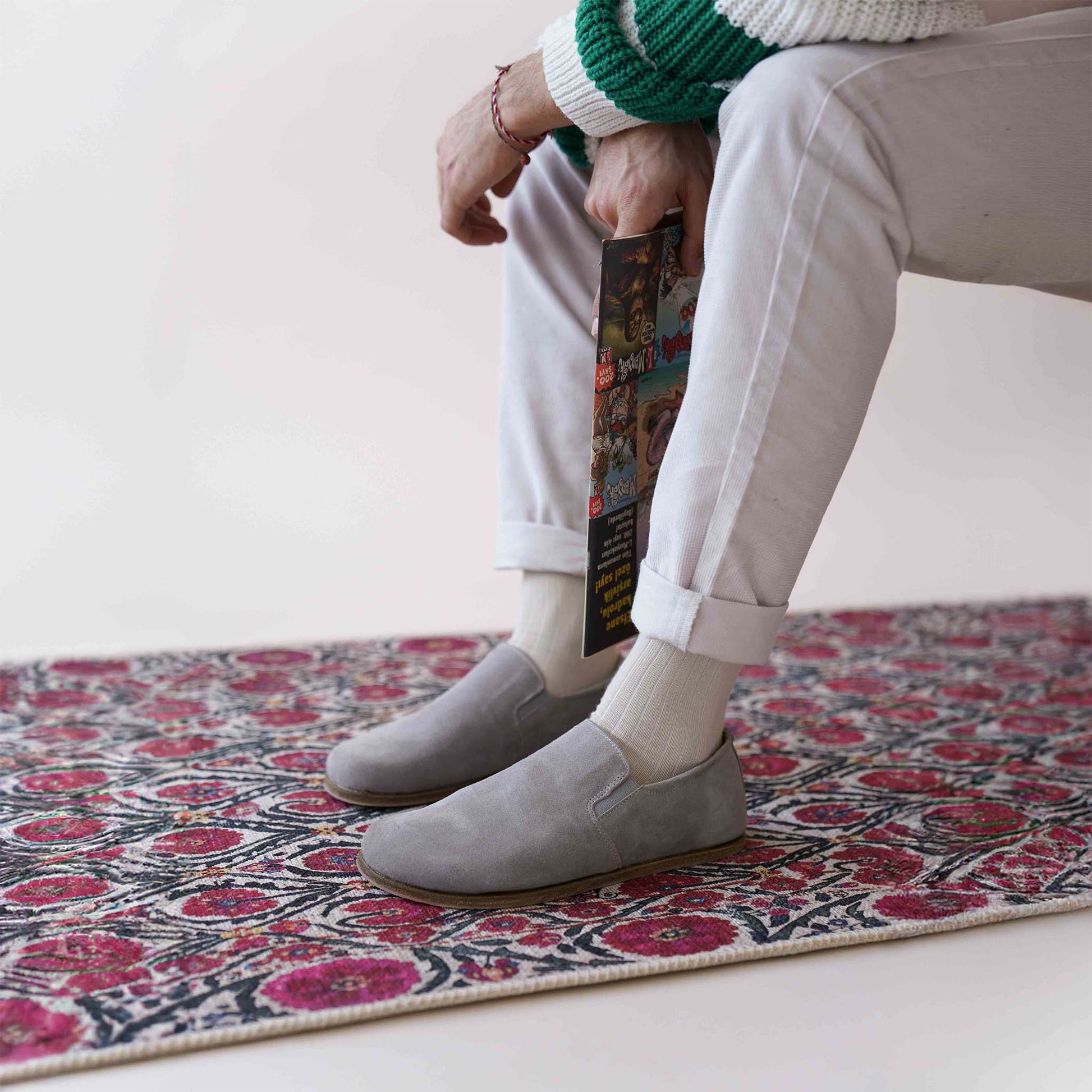 Model wearing gray suede barefoot men's loafers by Pelanir, sitting on a colorful rug with legs crossed.