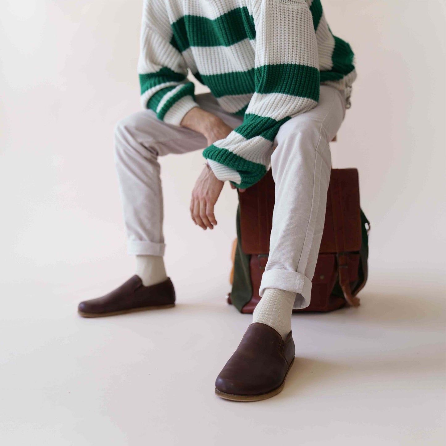 Man sitting with a backpack, wearing brown Ionia Leather Barefoot Men Loafers paired with white socks and light pants.