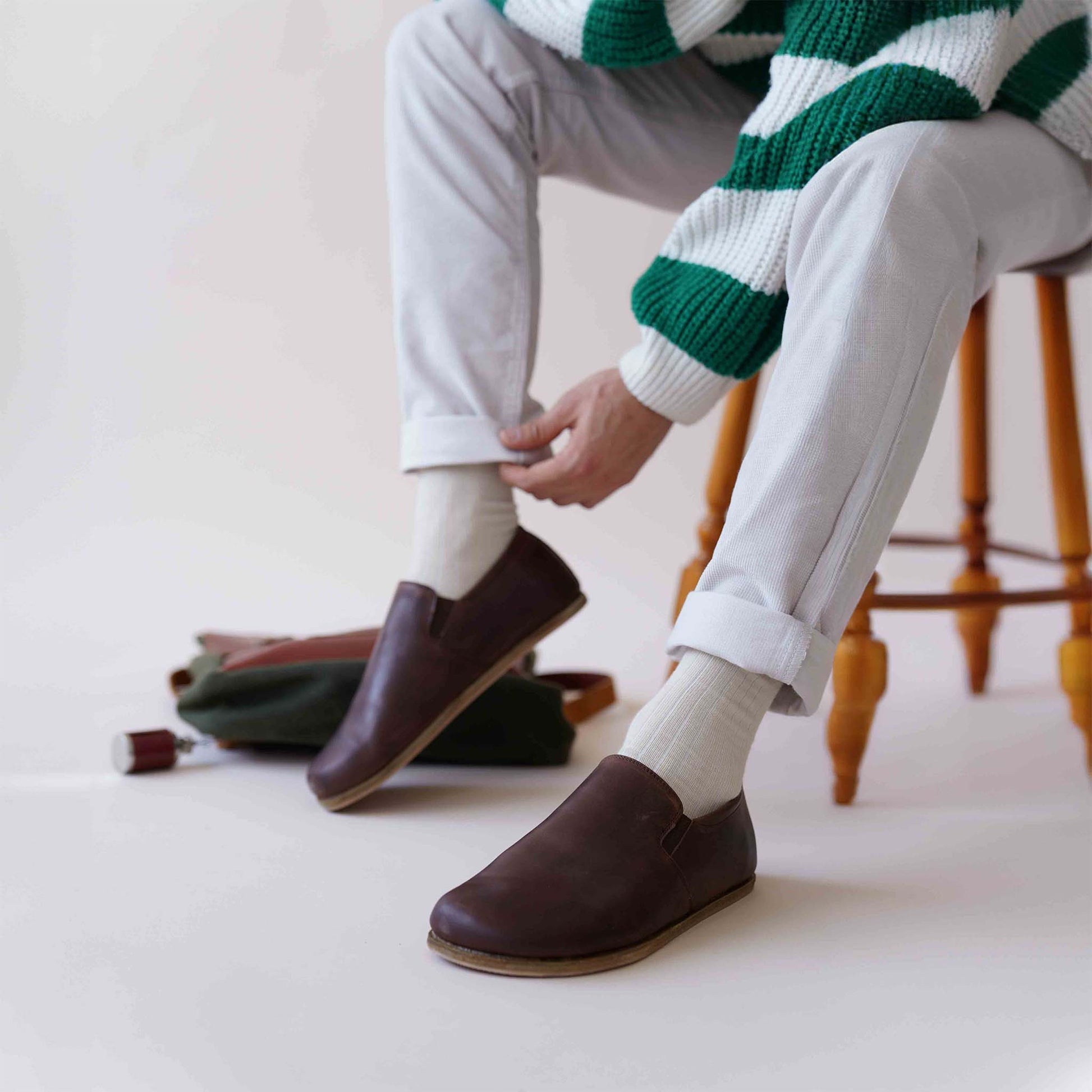 Person sitting with legs crossed, wearing brown Ionia Leather Barefoot Men Loafers, paired with white socks and light-colored pants.