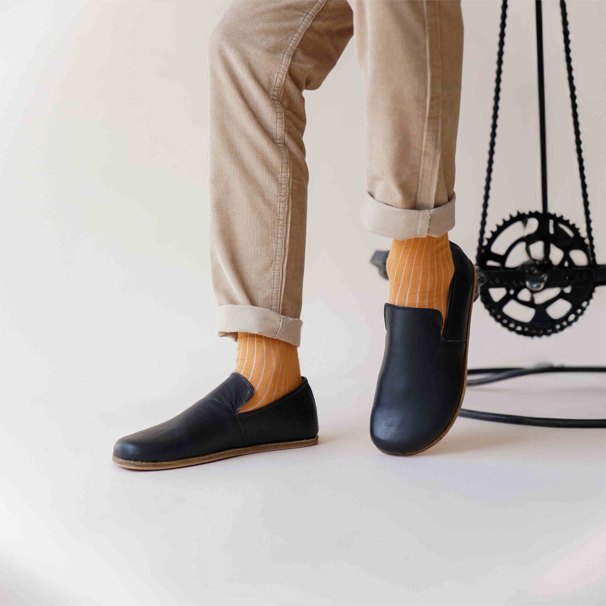 Man sitting on stool wearing Aeolia Leather Barefoot Men Loafers in black with beige pants and orange socks - shop at pelanir.com.