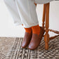 Model wearing Aeolia tan brown leather barefoot women loafers, paired with white pants and orange socks, seated on a wooden chair.