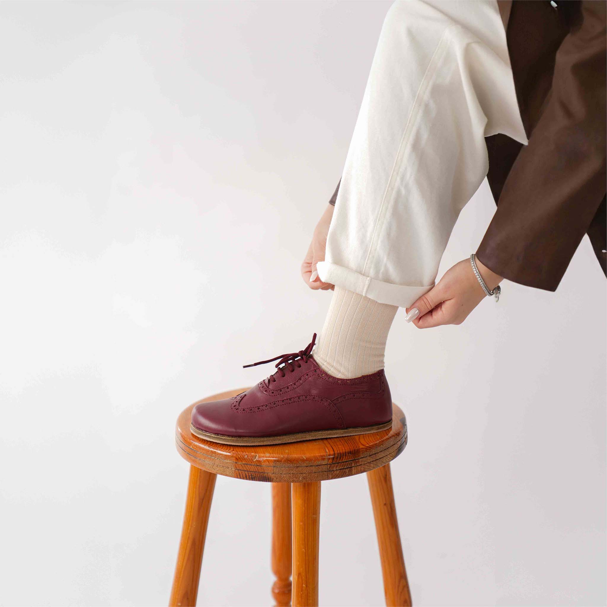 Close-up of a person adjusting white pants while wearing Doris leather barefoot women Oxfords in Burgundy, standing on a wooden stool.